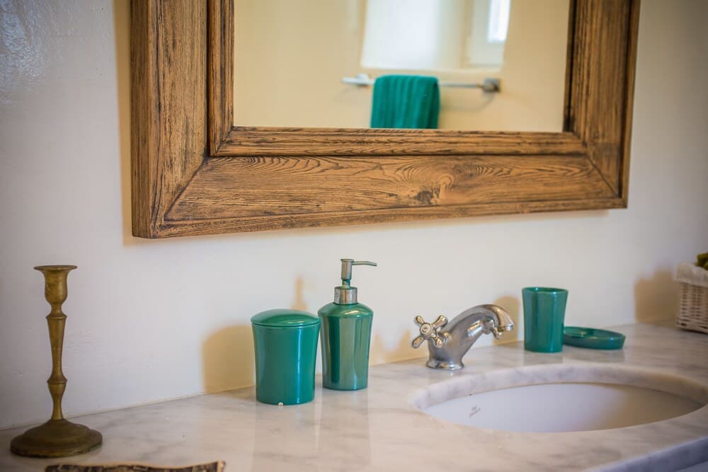 A cozy bathroom vanity features a rustic wooden-framed mirror above a marble countertop adorned with teal accessories and a brass candlestick, ideal for travelers seeking a serene and stylish retreat.
