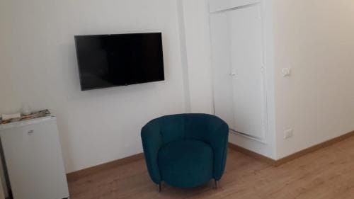 A cozy hotel room features a teal armchair, a mounted flat-screen TV, and a small fridge, creating a minimalist and inviting atmosphere ideal for travelers seeking comfort and simplicity.