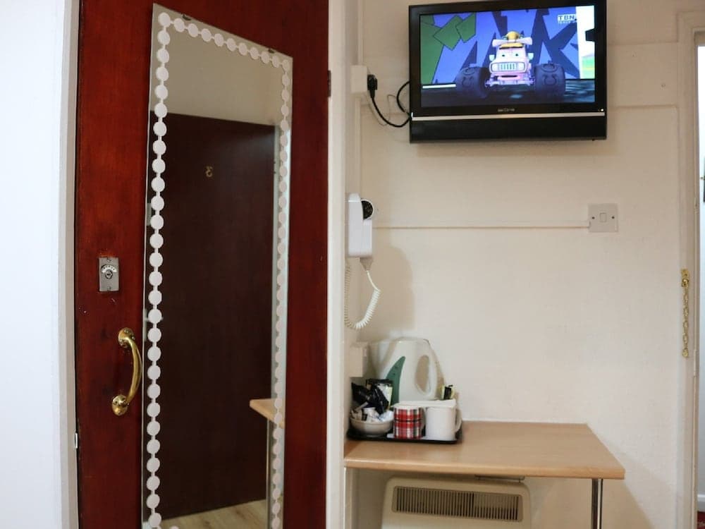 A cozy hotel room corner features a mounted TV playing a cartoon, a small desk with a kettle and mugs, and a decorative mirror beside the door, ideal for travelers seeking comfort and convenience.
