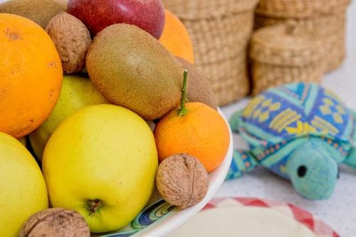 A vibrant bowl of fresh fruits like apples, oranges, kiwis, and walnuts sits beside a colorful turtle-shaped textile on a cozy tablecloth, perfect for a culinary-themed travel blog showcasing local markets or homey dining experiences.