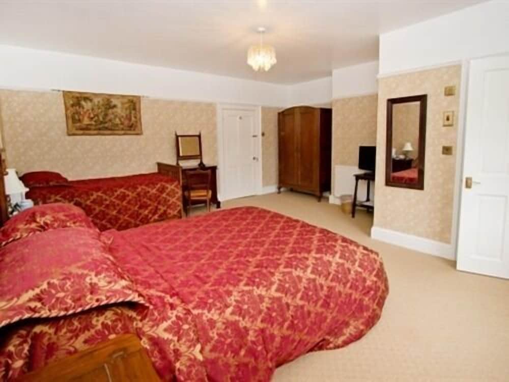 A cozy hotel room features two beds with red patterned quilts, a wooden wardrobe, a mirror, and warm lighting, ideal for travelers seeking comfort and a classic ambiance.