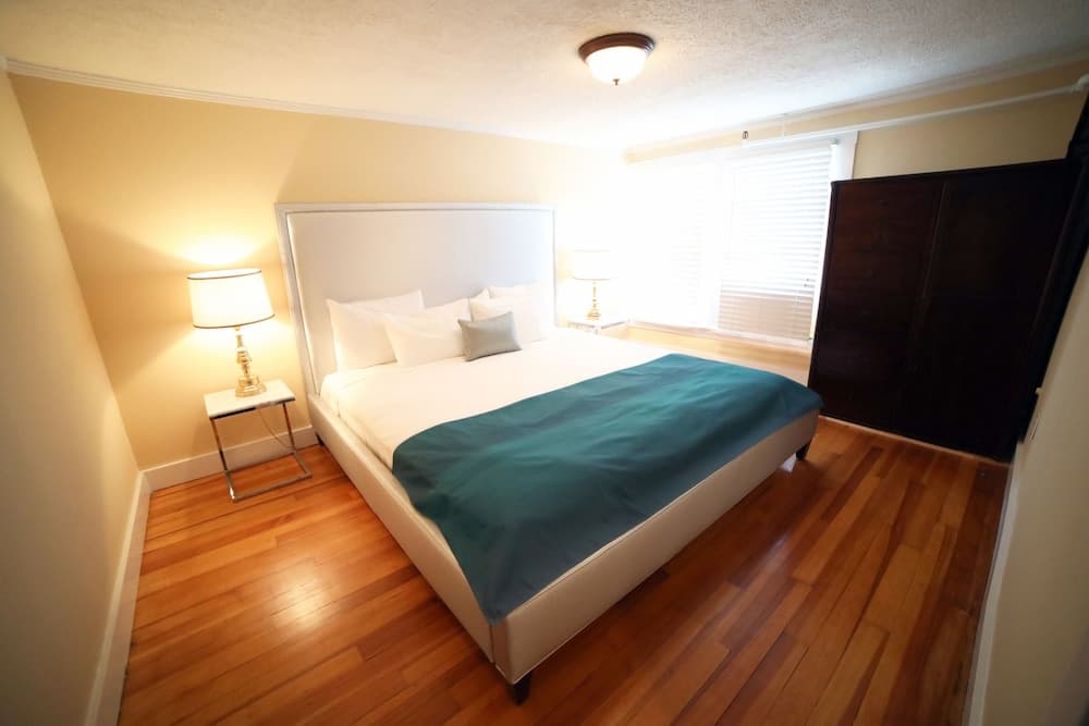 A cozy bedroom with hardwood floors, a neatly made bed adorned with a teal throw, warm bedside lamps, and natural light filtering through blinds, ideal for travelers seeking a comfortable and inviting accommodation.