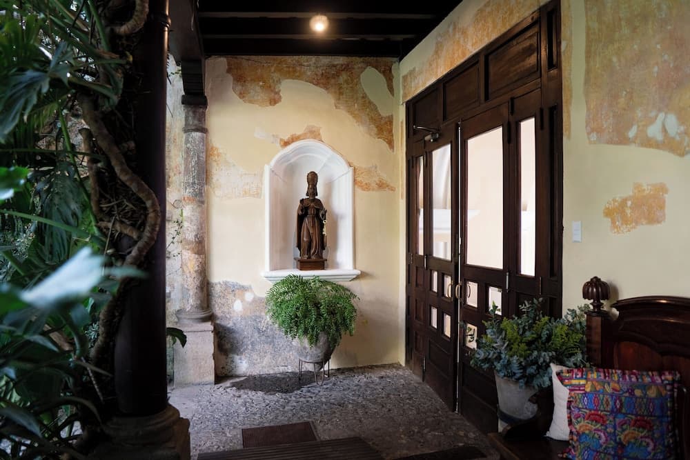 A serene indoor courtyard features rustic walls, a statue in an arched niche, lush greenery, and wooden doors, ideal for travelers seeking a tranquil historical ambiance.