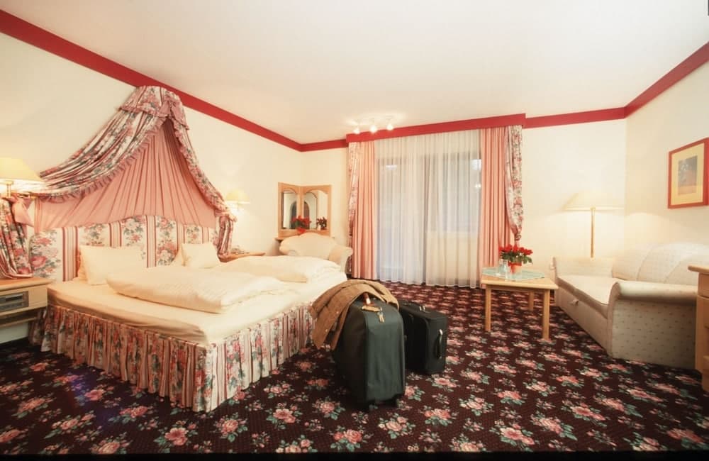 A cozy hotel room features a floral-patterned bed with a canopy, a floral carpet, and a seating area by the window, creating a warm and inviting atmosphere ideal for travelers seeking comfort and relaxation.