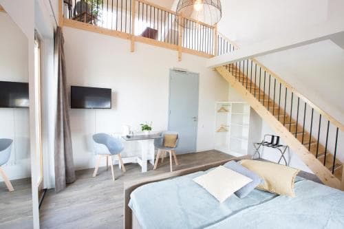 A cozy loft-style apartment features a modern bedroom with a mezzanine level, wooden staircase, and minimalist decor, ideal for travelers seeking a stylish and comfortable urban retreat.