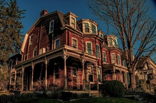 A grand Victorian-style brick house with intricate woodwork and a wraparound porch stands under a clear blue sky, evoking a nostalgic and serene mood, perfect for history enthusiasts exploring architectural landmarks.