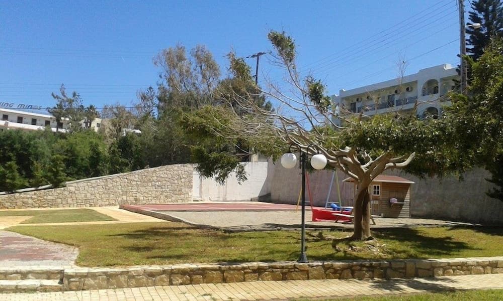 A serene park area with lush greenery, a stone wall, a swing set under a tree, and modern buildings in the background offers a peaceful retreat for travelers seeking relaxation and leisure activities.