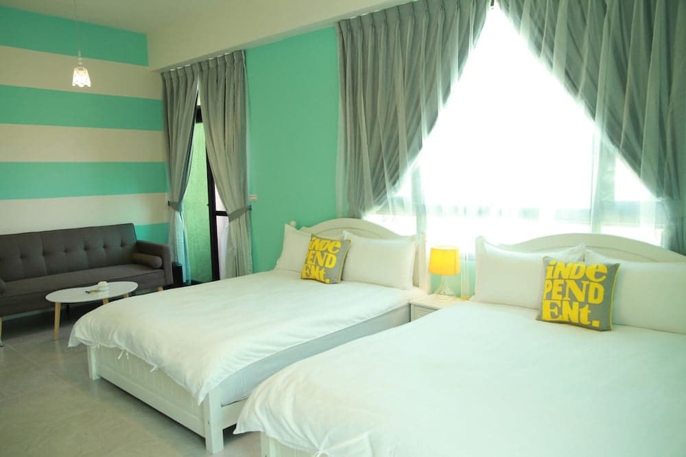 A cozy hotel room with twin beds adorned with "INDEPENDENT" pillows, turquoise-striped walls, a gray sofa, and large windows draped in sheer curtains, ideal for travelers seeking comfort and style.