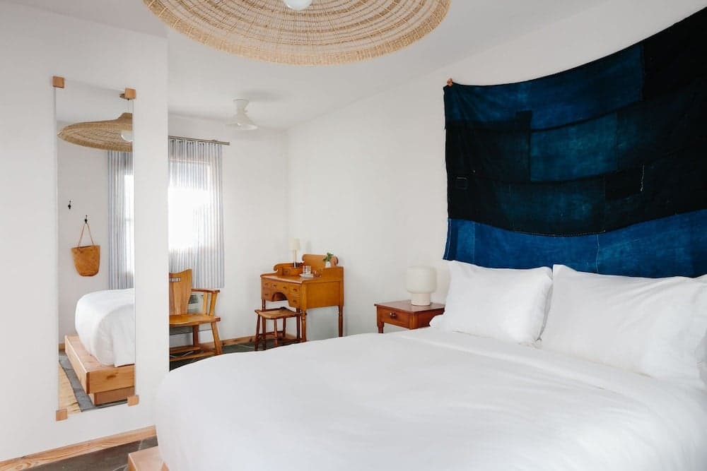 A serene hotel bedroom features a cozy bed adorned with white linens, a blue tapestry wall hanging, wooden furniture, and a woven ceiling lamp, ideal for travelers seeking a tranquil retreat.