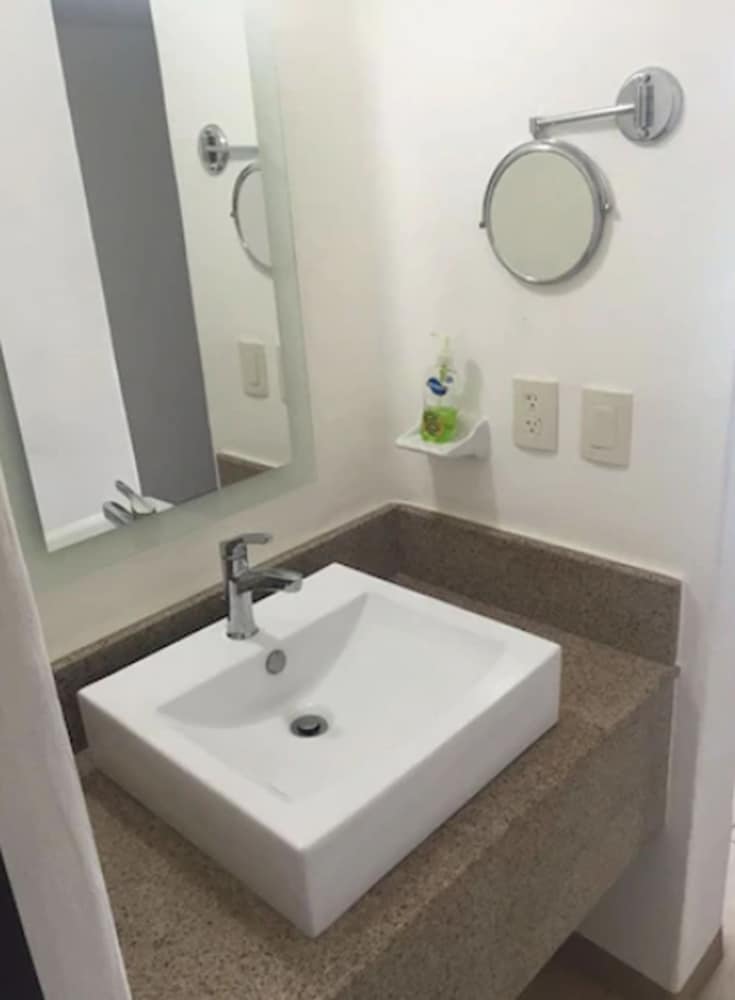 A modern bathroom features a sleek white sink on a granite countertop, a wall-mounted magnifying mirror, and a minimalist design, ideal for travelers seeking clean and functional accommodations.