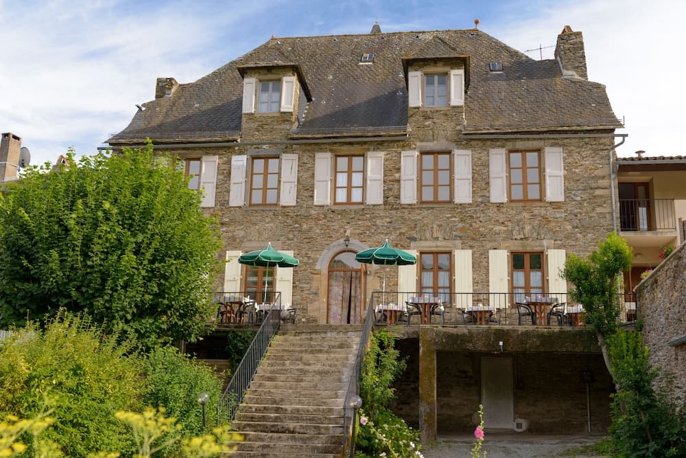 A charming stone building with white shutters and green umbrellas invites travelers to relax in its quaint outdoor seating area, perfect for a serene European getaway.