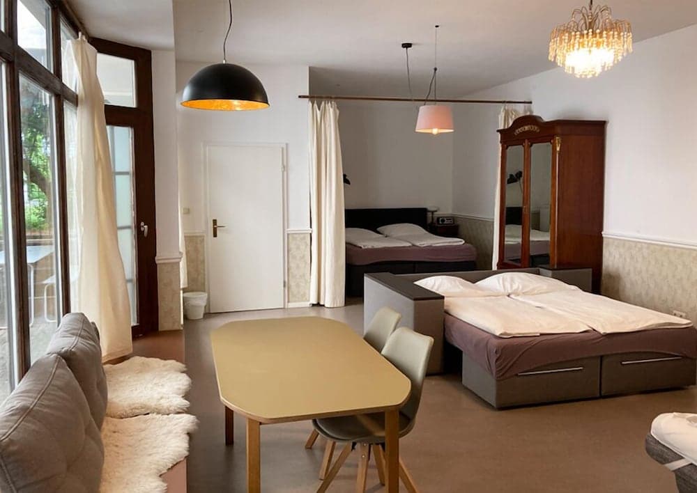 A cozy studio apartment features a modern dining area, two neatly made beds, a vintage armoire, and large windows with a serene view, ideal for travelers seeking comfort and style in a compact space.