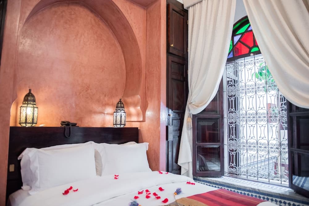 A cozy, warmly-lit bedroom with an arched alcove, ornate lanterns, and a stained-glass window framed by flowing curtains invites travelers seeking a romantic and culturally rich retreat.