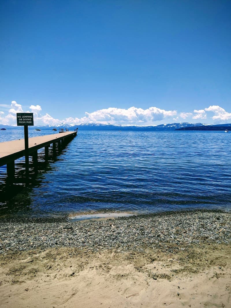 A serene lakeside scene at the Franciscan Lakeside Lodge features a long wooden pier extending into calm blue waters under a clear sky, with a "No Jumping or Diving" sign, ideal for tranquil vacations and nature photography.