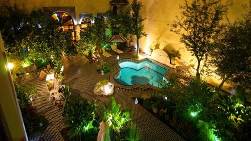 A serene courtyard hotel scene illuminated at night features a heart-shaped pool surrounded by lush greenery and cozy seating areas, ideal for romantic getaways or tranquil retreats.