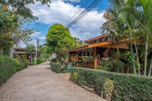 A serene tropical pathway lined with lush greenery leads to a charming wooden restaurant under a partly cloudy sky, ideal for travelers seeking a peaceful dining experience in a natural setting.