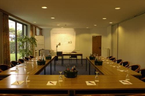 A well-lit conference room with wooden tables set for a meeting, featuring a whiteboard, plants, and large windows overlooking greenery, ideal for professional gatherings and team discussions.