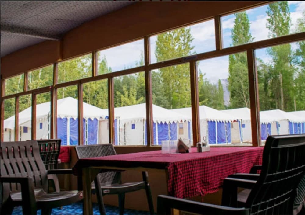 A cozy indoor dining area with large windows offers a serene view of lush green trees and white tents outside, ideal for travelers seeking a tranquil camping retreat experience.