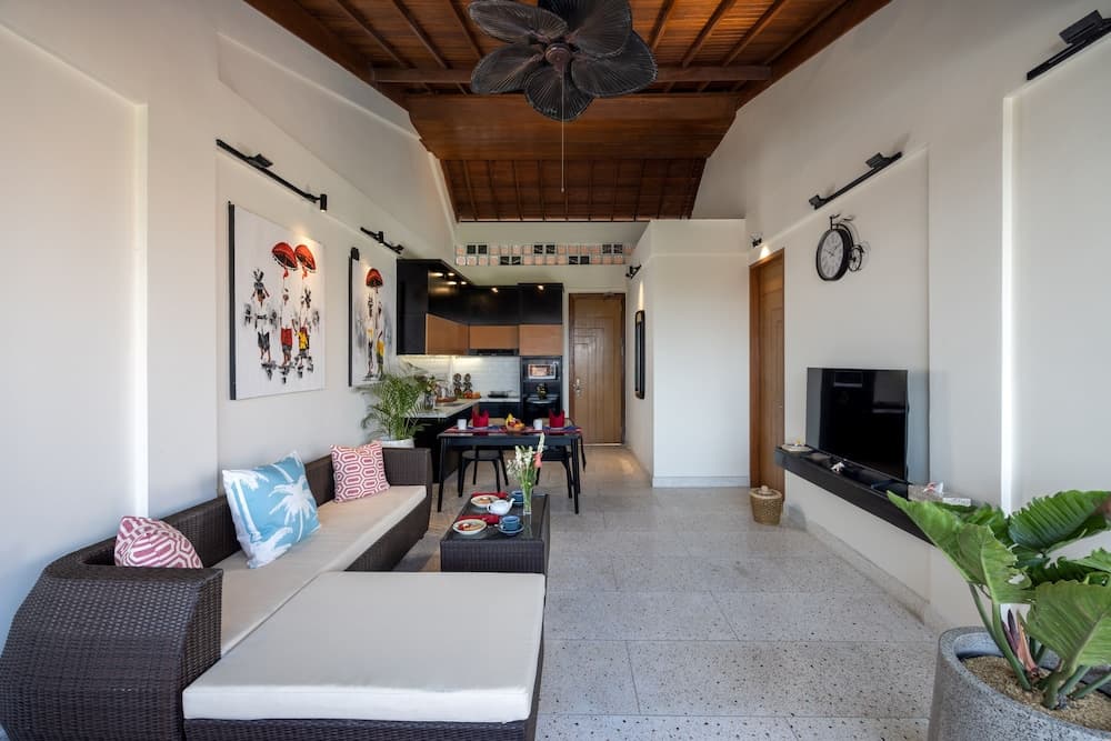 A cozy tropical villa living space features a wicker sofa adorned with colorful cushions, a sleek kitchen in the background, and vibrant greenery, ideal for travelers seeking a serene and stylish retreat.