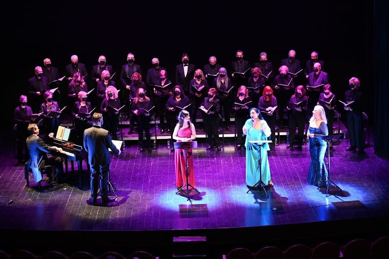A vibrant concert scene at an indoor theater features a choir in black attire, three soloists in colorful dresses, and a pianist, all illuminated by purple stage lighting, creating an enchanting atmosphere ideal for cultural and musical exploration.