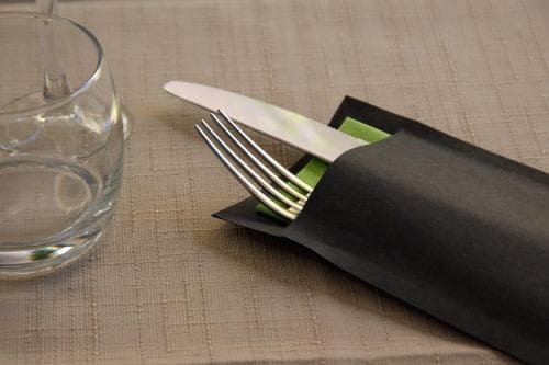 A neatly arranged dining table setting featuring a glass of water, cutlery wrapped in a black napkin with a green accent, evokes a sophisticated dining experience ideal for travelers seeking upscale restaurant ambiance.