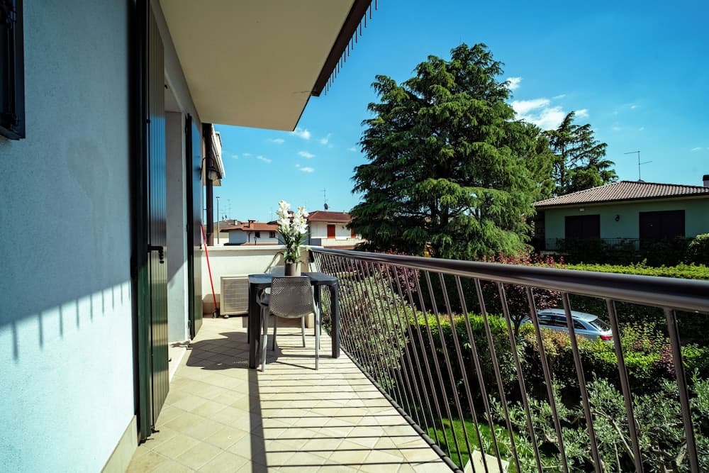 A serene balcony with a tiled floor, metal railing, and a small table set with flowers overlooks lush greenery and residential rooftops under a clear blue sky, ideal for travelers seeking a peaceful retreat.