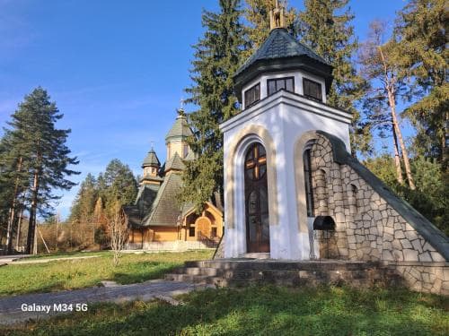 A serene forest setting features a charming stone chapel with wooden accents and a traditional church in the background, ideal for spiritual retreats and nature lovers exploring religious heritage sites.