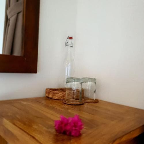A serene minimalist room corner features a wooden table adorned with glassware, a woven basket, and vibrant pink flowers, ideal for travelers seeking a tranquil and stylish accommodation setting.