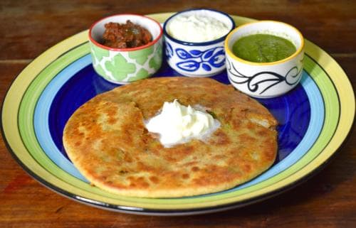 A vibrant plate of stuffed flatbread served with dollops of yogurt and colorful chutneys on a rustic wooden table, perfect for exploring local culinary delights during a food-focused travel adventure.