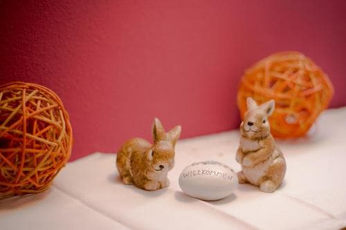 A cozy indoor setting features charming ceramic bunnies beside an egg inscribed with "WIEKOMMEN," flanked by decorative woven balls, ideal for Easter-themed travel inspiration or home decor ideas.