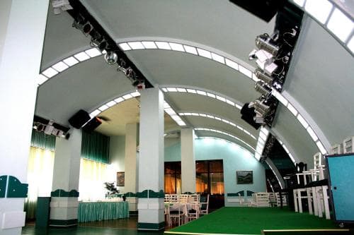 A modern event hall with arched ceilings, green carpeting, and stage lighting creates an inviting atmosphere ideal for conferences or gatherings, featuring tables, chairs, and decorative elements for versatile travel arrangements.
