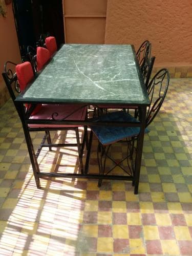A cozy outdoor dining area features a green marble table surrounded by colorful cushioned chairs on a vibrant checkered tile floor, perfect for travelers seeking a relaxed café experience.