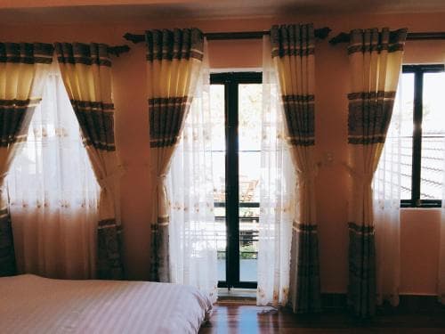 A cozy hotel room with soft lighting, patterned curtains framing large windows, and a neatly made bed invites travelers seeking comfort and relaxation.