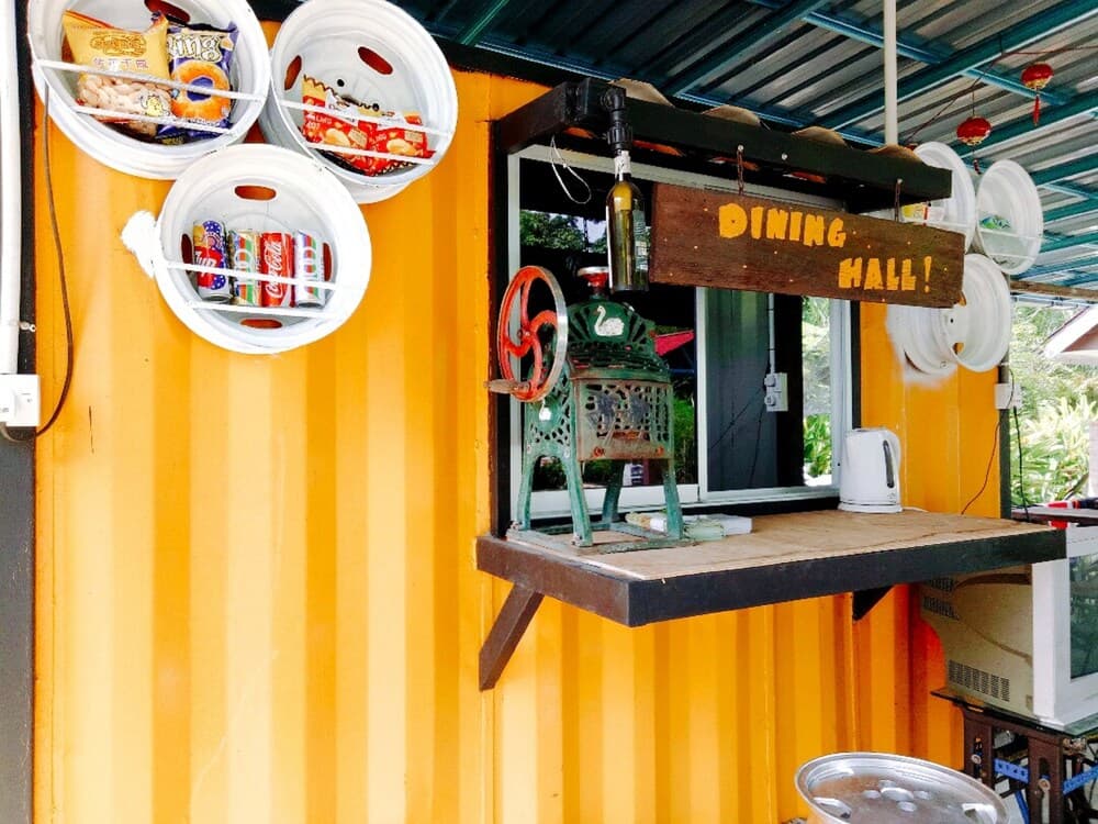 A vibrant yellow shipping container hosts a quirky outdoor dining setup with a "Dining Hall!" sign, colorful drink buckets, and eclectic decor, perfect for travelers seeking unique and cozy eating spots.
