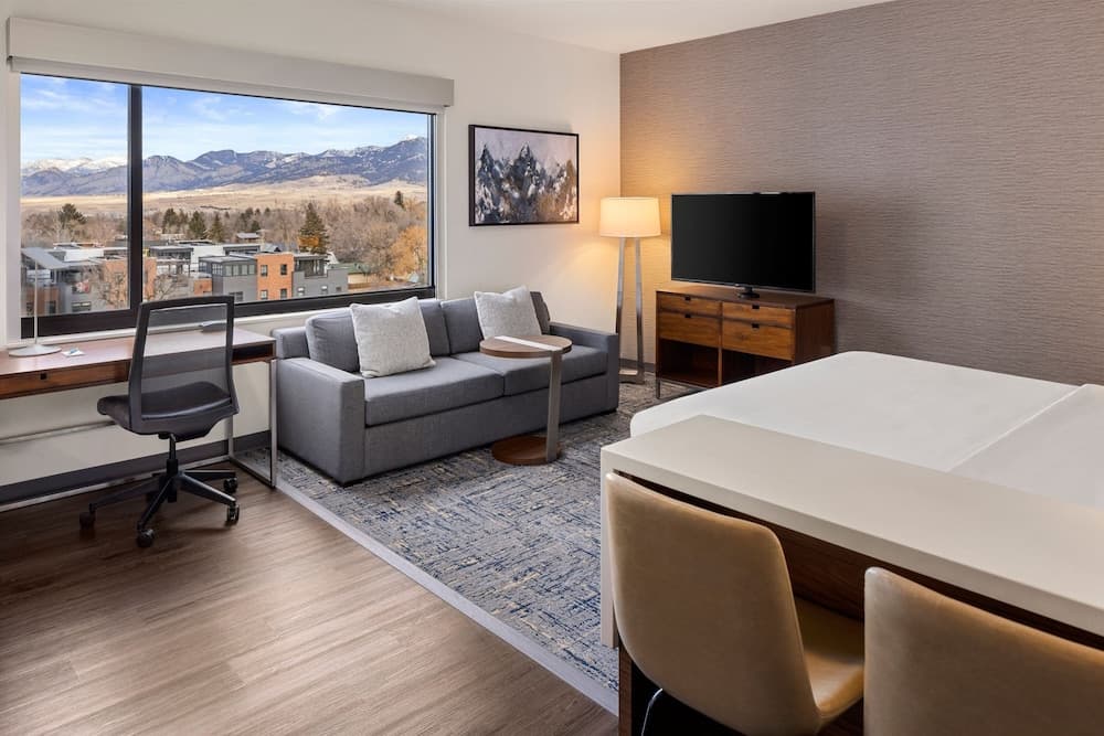 A cozy hotel suite with a scenic mountain view through large windows, featuring modern furnishings like a sofa, desk, and dining area, ideal for travelers seeking comfort and productivity in a serene setting.