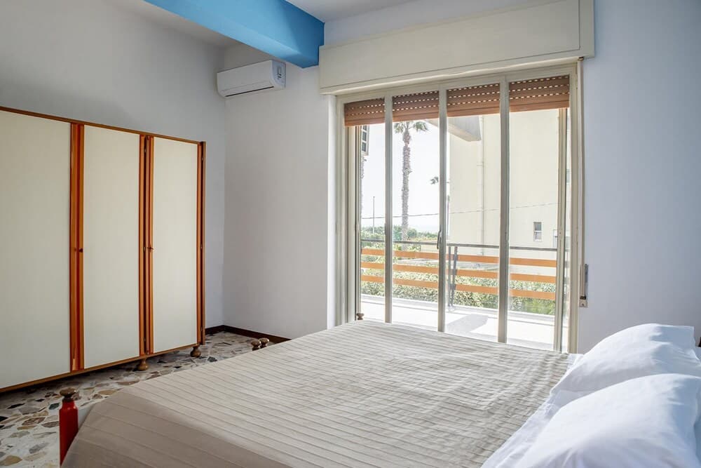 A serene hotel bedroom with a neatly made bed, wooden closet doors, and a sliding glass door leading to a balcony with palm trees, ideal for travelers seeking a relaxing retreat.