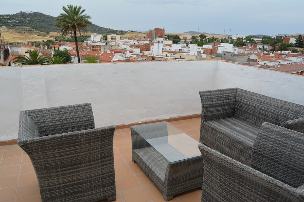 A serene rooftop terrace with wicker furniture overlooks a picturesque town nestled among rolling hills, ideal for travelers seeking relaxation and scenic views.