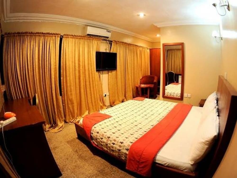 A cozy hotel room features a neatly made bed with red accents, golden curtains, a mounted TV, and warm lighting, ideal for travelers seeking comfort and relaxation.