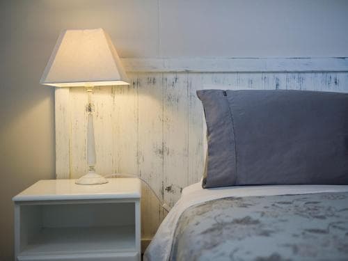 A cozy bedroom with a softly lit lamp on a white bedside table beside a bed with gray pillows, featuring rustic wooden paneling, ideal for travelers seeking a serene and comfortable stay.