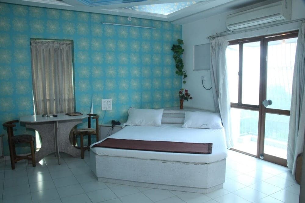 A cozy hotel room features a neatly made bed, a small desk by a window with blue floral wallpaper, and a sliding glass door leading to natural light, ideal for travelers seeking comfort and simplicity.