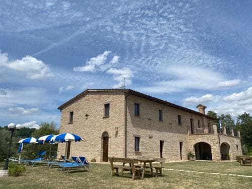 A serene countryside villa with rustic stone architecture, surrounded by lush greenery under a vibrant blue sky, offers a tranquil retreat complete with outdoor seating and umbrellas, ideal for a relaxing vacation getaway.