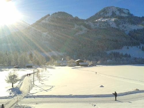 A serene winter landscape features snow-covered mountains, a frozen lake with skaters, and quaint cabins under a radiant sun, ideal for a peaceful skiing getaway.