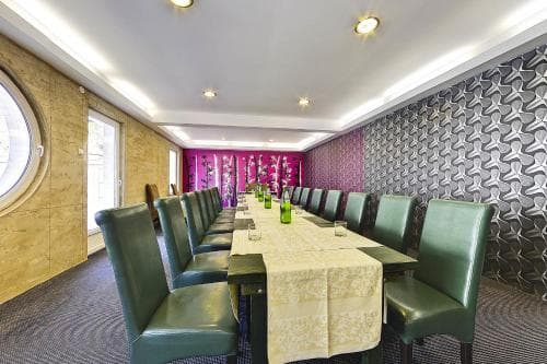 A cozy dining room with green chairs, a long table adorned with a yellow tablecloth and green bottles, set against patterned walls and a round window, evokes a warm and inviting atmosphere ideal for intimate gatherings or small events.