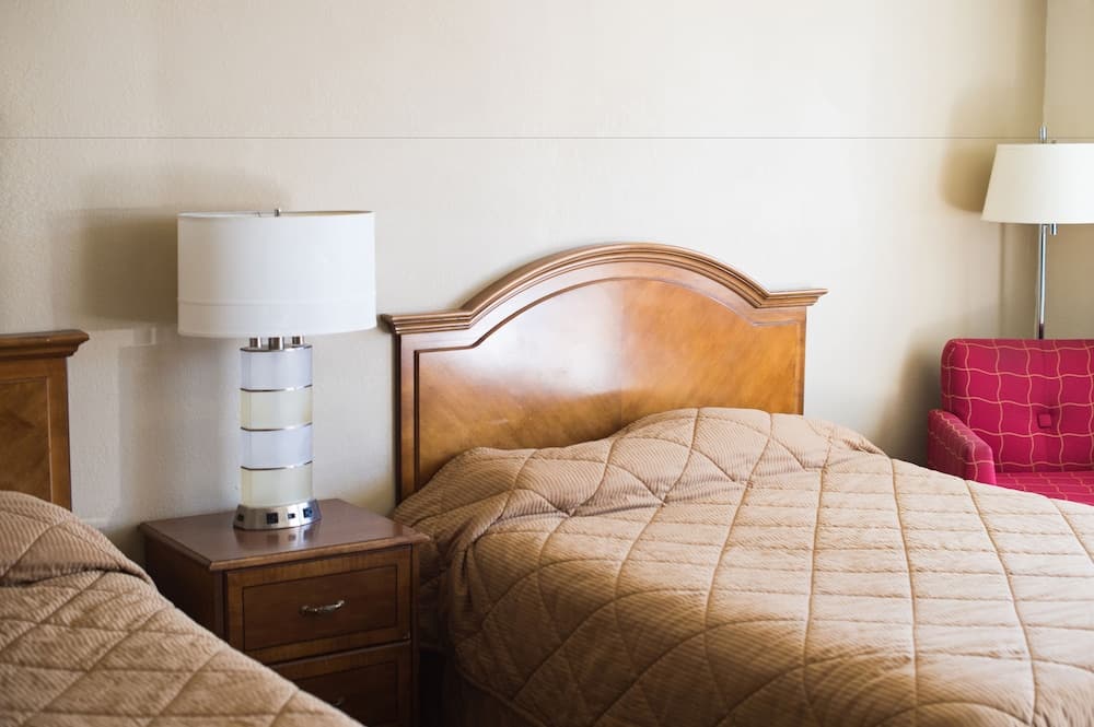 A cozy hotel room features two neatly made beds with quilted beige bedding, wooden headboards, bedside lamps, and a vibrant pink armchair, ideal for travelers seeking comfort and relaxation.