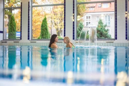 Two individuals relax in a serene indoor pool illuminated by natural light from large windows, featuring a decorative waterfall and surrounded by autumnal trees, ideal for a tranquil spa getaway.