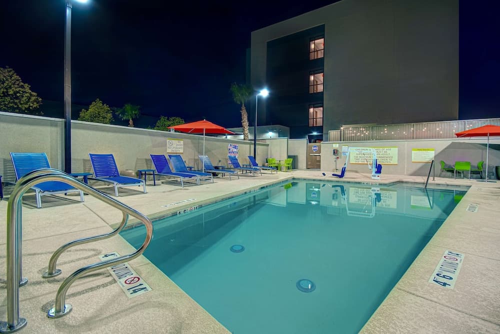 A serene nighttime scene at an outdoor hotel pool area, featuring illuminated blue lounge chairs, red umbrellas, and palm trees, ideal for travelers seeking relaxation during evening hours.