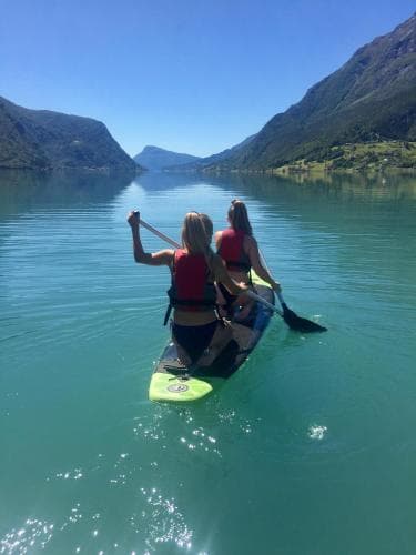 Two adventurers paddleboard across a serene turquoise lake surrounded by towering mountains under a clear blue sky, capturing an idyllic outdoor activity scene perfect for adventure seekers.