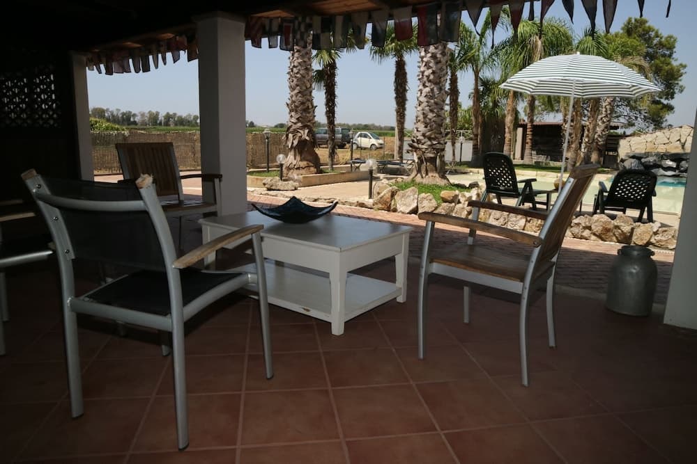 A serene outdoor patio shaded by a pergola features comfortable seating around a table, surrounded by palm trees and a tranquil poolside ambiance, ideal for relaxing vacations or leisurely getaways.