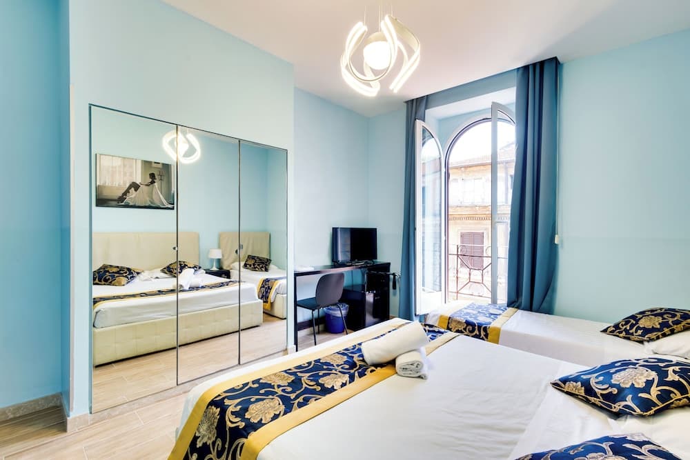 A serene hotel room with light blue walls, twin beds adorned with blue and gold bedding, a mirrored wardrobe, and an arched window offering a view of traditional architecture, ideal for travelers seeking comfort and charm.