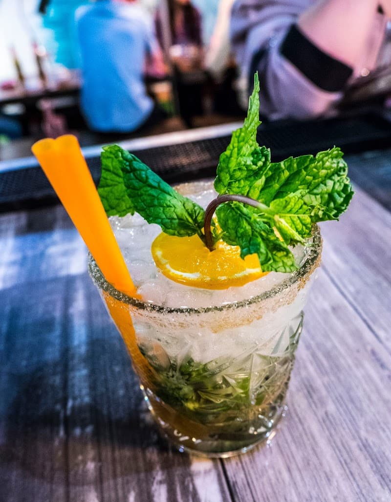 A refreshing mojito garnished with mint and orange sits invitingly on a wooden table at the cozy La Banda Rooftop Hostel in Sevilla's historic Casco Antiguo, perfect for travelers seeking a vibrant nightlife experience.